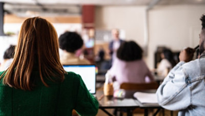 View from behind of a long-haired woman using a laptop in a classroom environment.