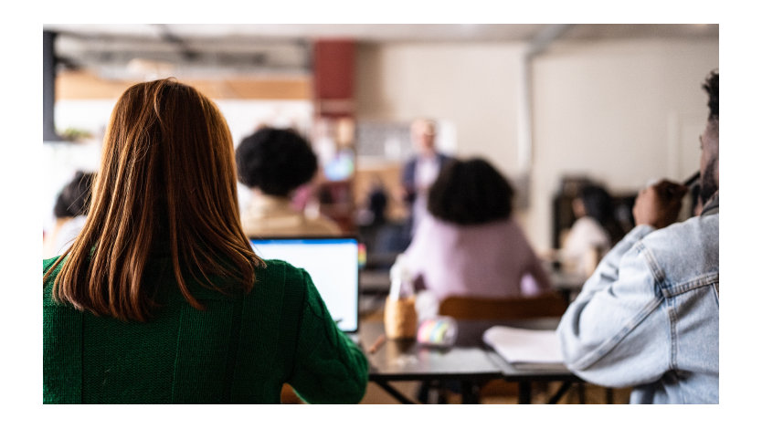 View from behind of a long-haired woman using a laptop in a classroom environment.