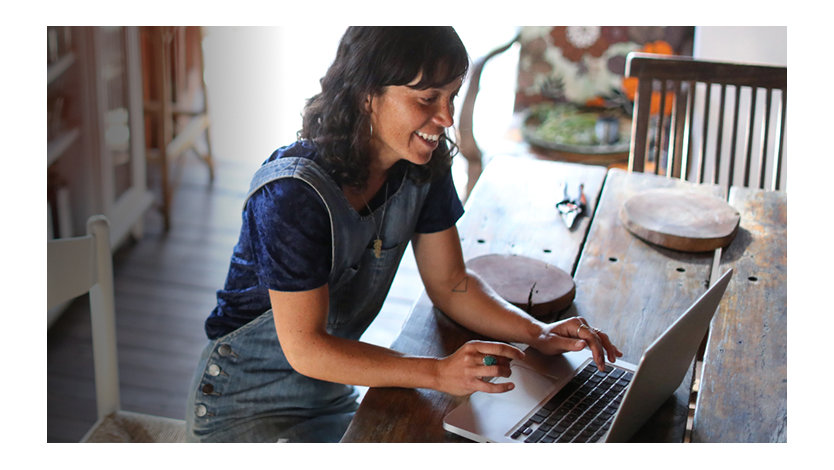 A smiling, casually-dressed woman works on a laptop in her home.