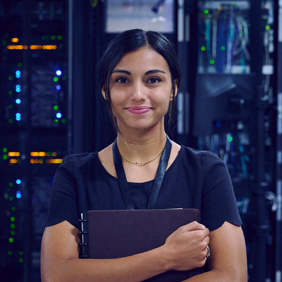 A female IT employee stands in a server room smiling and holding a folder.