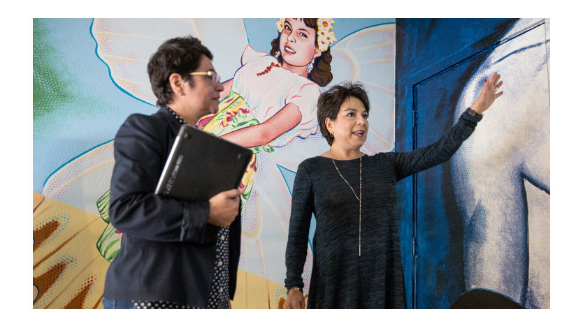Two people, one holding a laptop, standing in front of a decoratively painted wall.