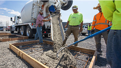 Construction workers pour concrete from a large mixing truck into a frame on the ground.