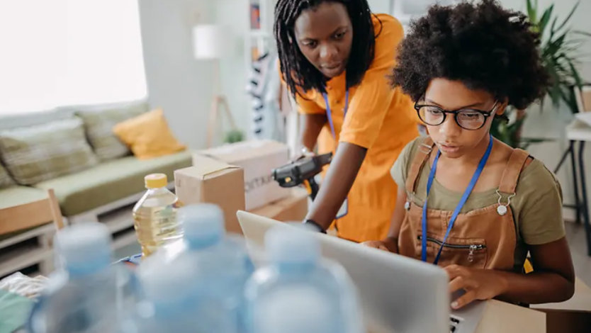 Two women working together on a laptop amid packaged goods and water bottles.