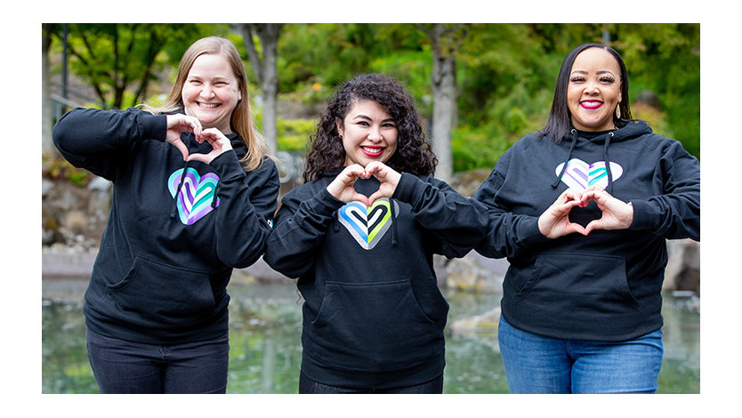Three volunteers wearing heart sweatshirts smiling and making heart gestures with their hands.