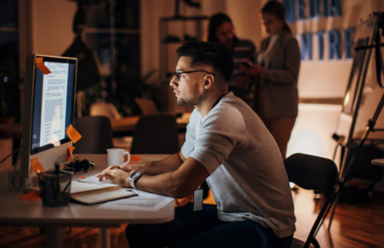 A man in an office writes a document that can be seen on his monitor, while two people with media equipment stand in the background.