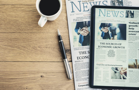 An overhead view of a desktop with a newspaper, tablet device displaying a news page, pen, and a cup of coffee.