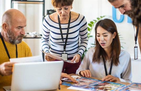 Five people in an office discuss various printed materials and magazines.