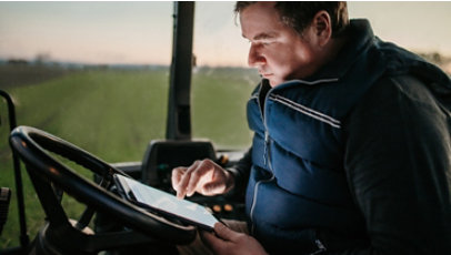 A man seated outdoors behind the wheel of a farming vehicle looks at a tablet device.