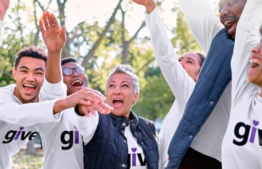 People outdoors in white “give” t-shirts celebrating with their hands in the air.