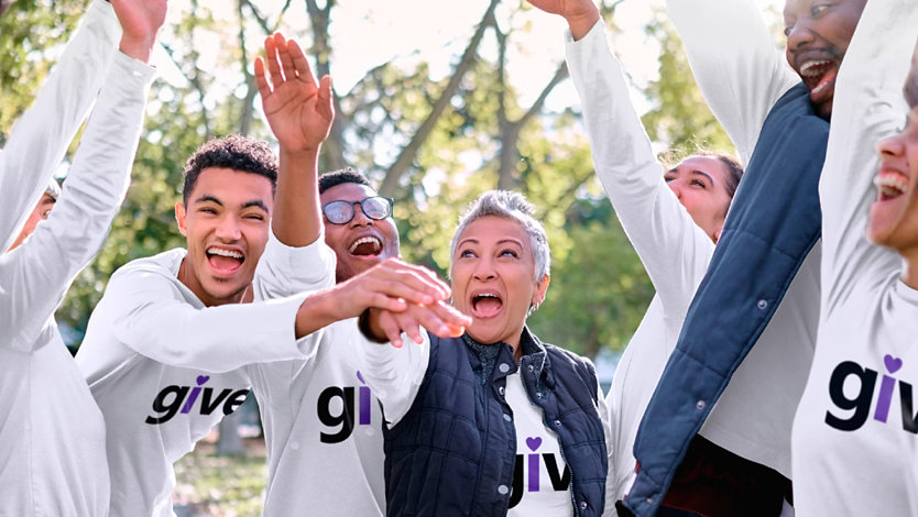 People outdoors in white 'give' t-shirts celebrating with their hands in the air.