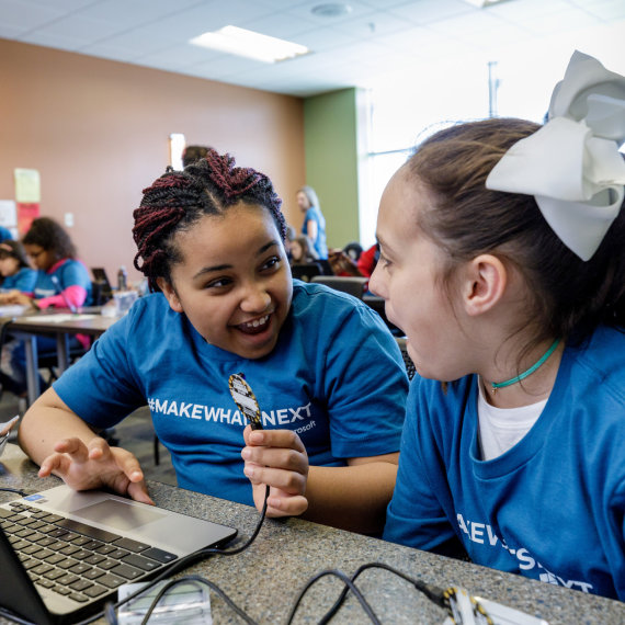 Two elementary school girls laugh while playing with a small device attached to a laptop