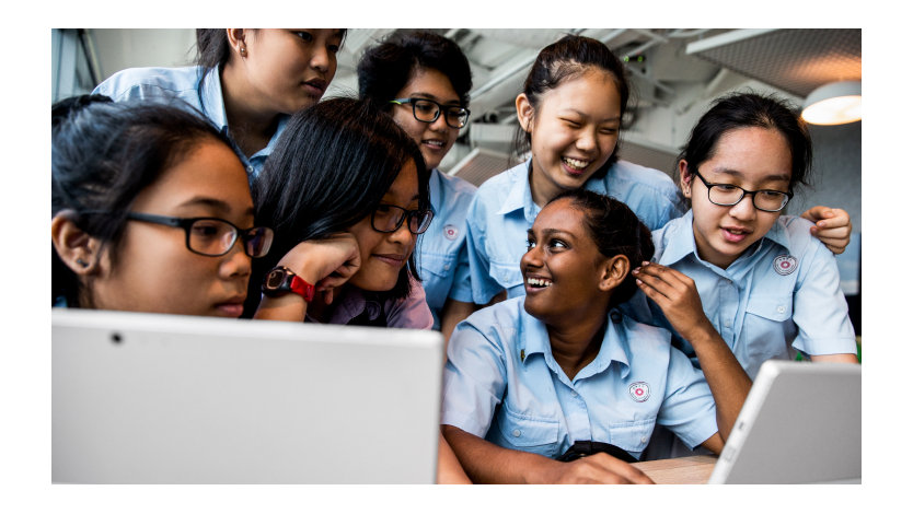 A group of young people in uniforms look at computer monitors together.