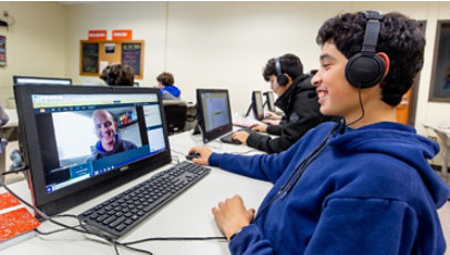 A young person wearing headphones in a computer lab smiles at a person on the computer screen in front.