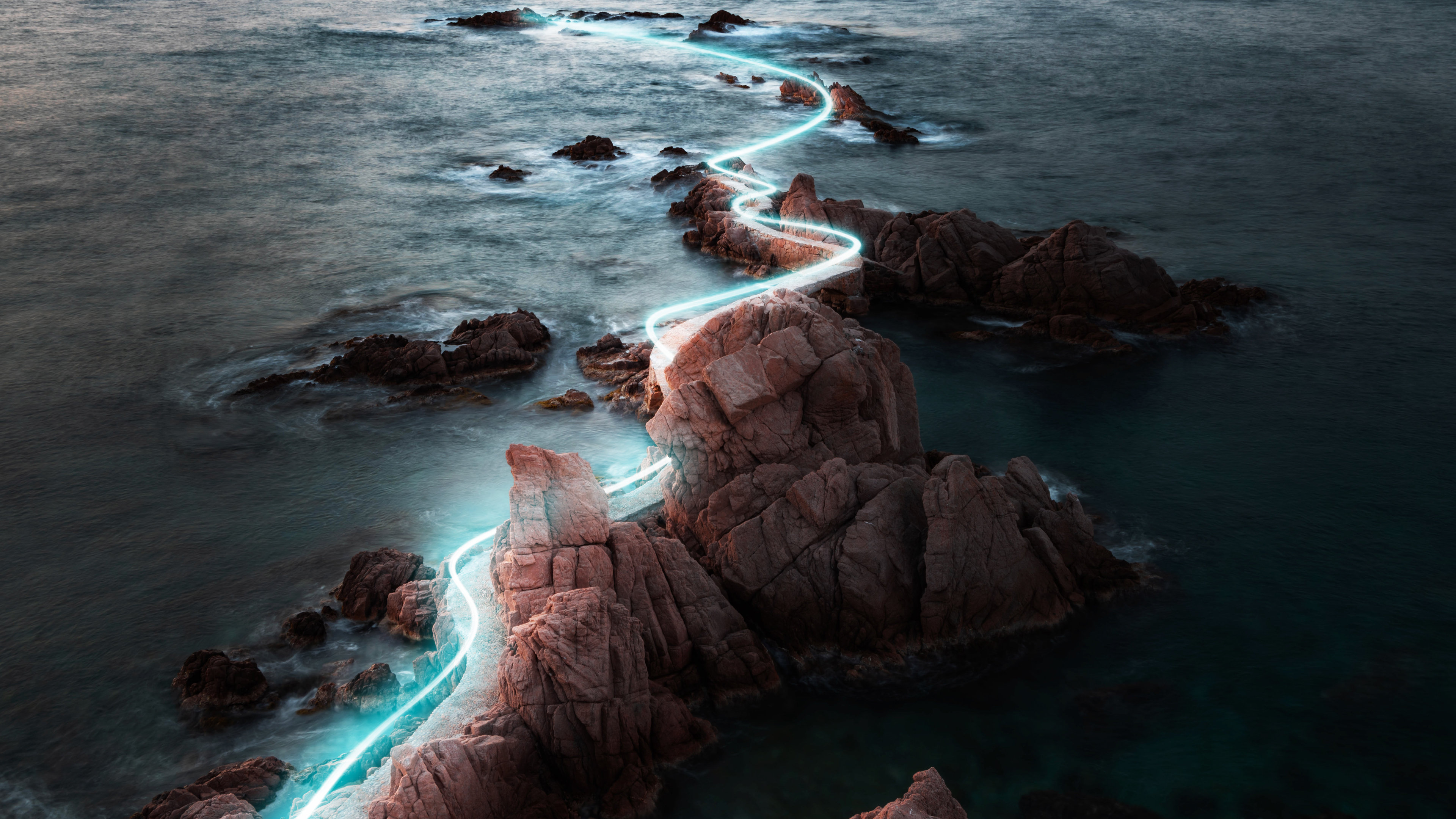 A glowing light trail winds across rocks in the ocean.