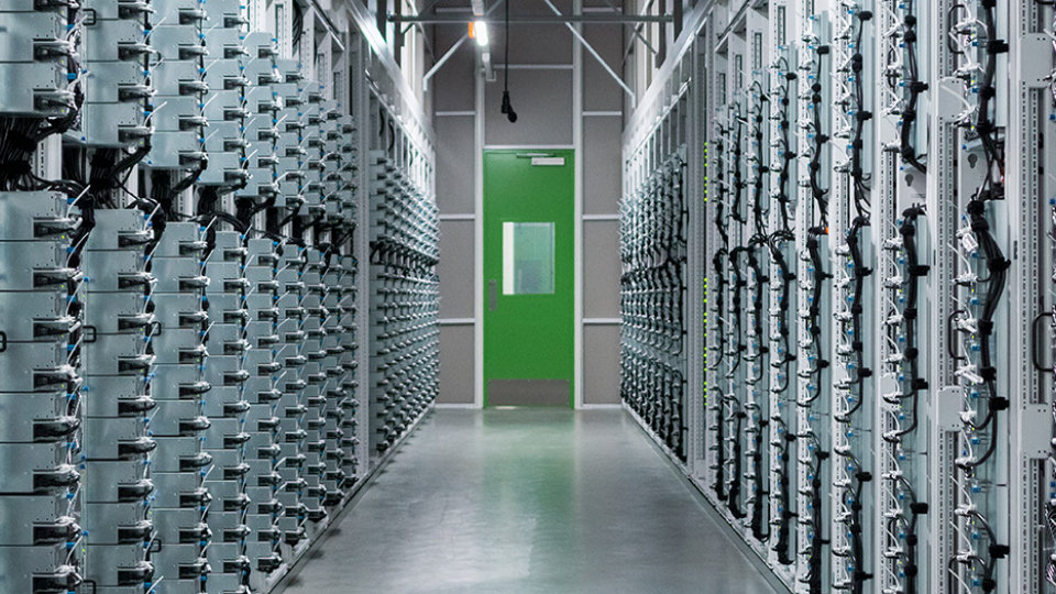 Aisle between rows of equipment racks in a data center, ending at a bright green door.