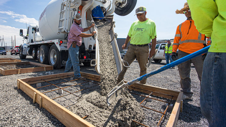 Construction workers pour concrete from a large mixing truck into a frame on the ground.