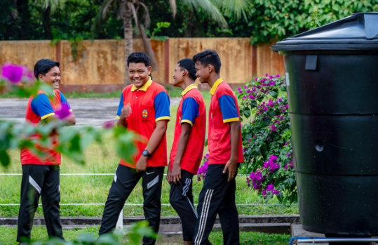 Four students in red shirts near a large water tank used for harvesting rainwater in Malaysia.