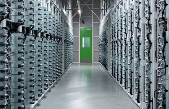 Long corridor of densely packed server racks in a datacenter, leading to a green door.