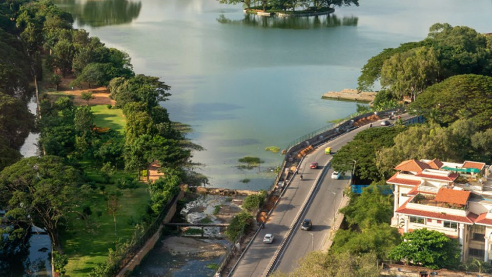 Aerial view of a city with a large lake and small island, a tree-lined road, and a distant skyline under a cloudy sky.