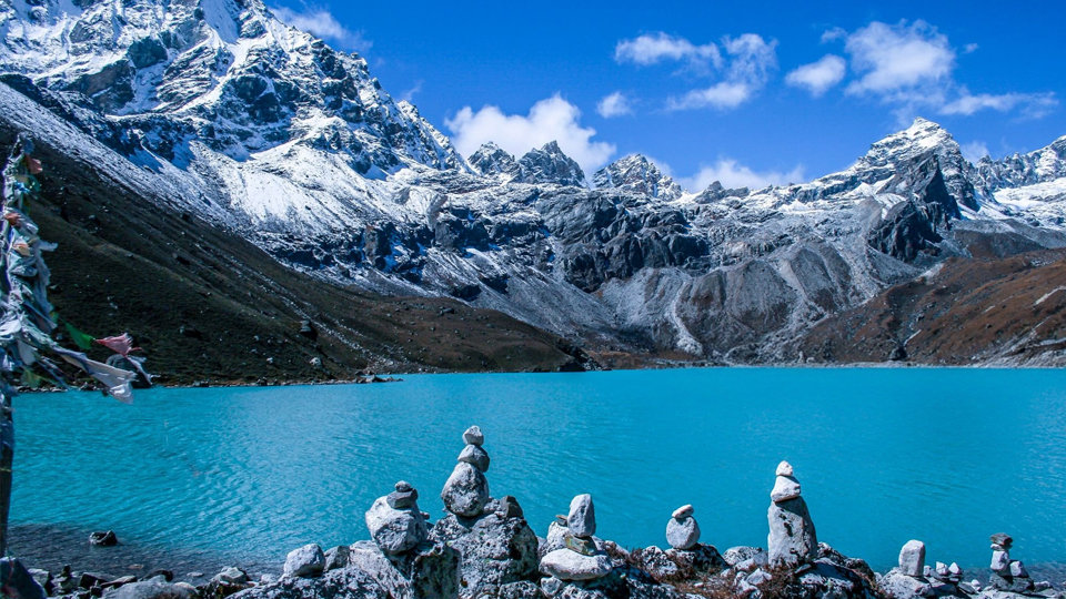 Turquoise glacial lake beneath snow-capped mountains with stone cairns in the foreground.