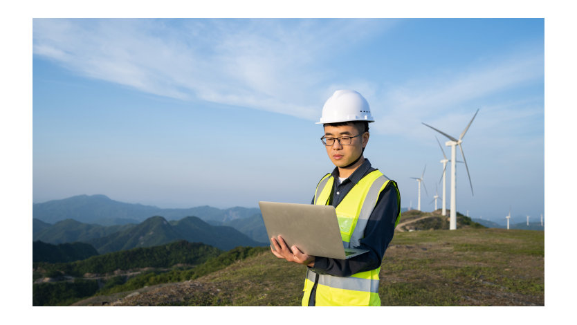 A man wearing a reflective vest and hardhat stands outside looking at a laptop with a mountainous landscape filled with wind turbines in the background.