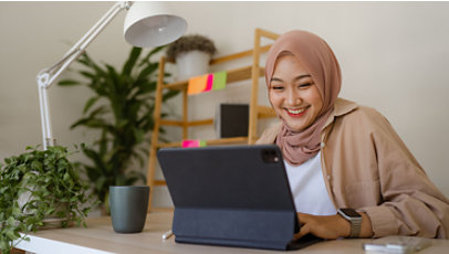 A woman wearing a hijab and surrounded by plants typing on her laptop at home.