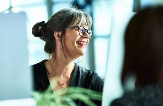 Close-up of a smiling woman in a business environment with computer monitors and plants in the foreground.