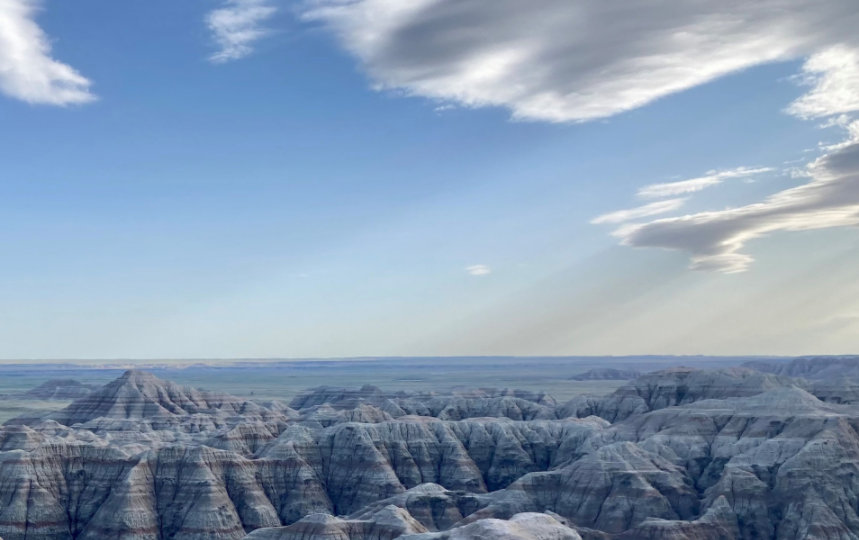 Landscape of mountains in the Badlands National Park with distinguished bands of color throughout the formations, blue skies with some clouds present. 