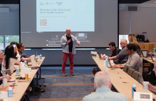 A speaker stands before a projection screen while participants at long tables work on laptops.