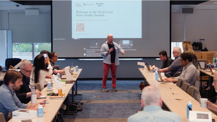A speaker stands before a projection screen while participants at long tables work on laptops.