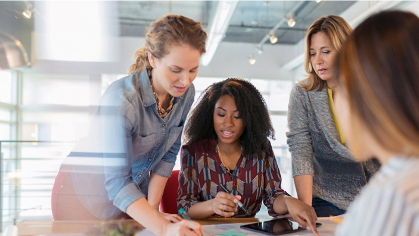 A diverse group of businesswomen look at a digital tablet and discuss a project in their modern office.