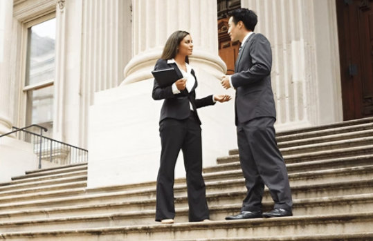 Two business professionals, a woman and a man, stand on the wide stone steps of a courthouse building with large white columns. They are engaged in a conversation; the woman holds a black portfolio and gestures with her hand, while the man listens attentively.