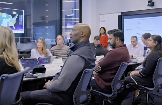 Election night meeting of Microsoft’s Election Situation Room, showing staff seated at a conference table looking at a speaker to the left with large wall-mounted screens in the background displaying election data and dashboards.