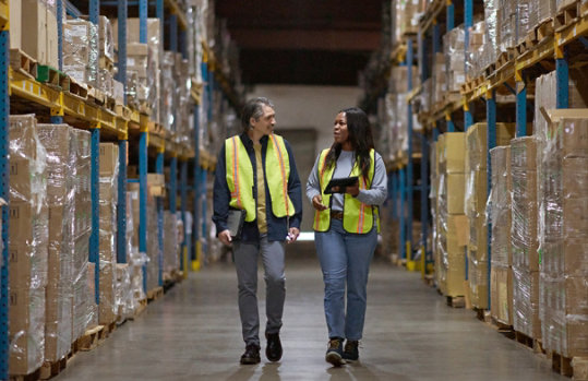 An image of two warehouse workers, a man and a woman, walking and talking in a large warehouse aisle. They are both wearing bright yellow safety vests. The woman is holding a tablet computer. The aisle is flanked by tall metal racks filled with boxes and packages stacked on pallets. The warehouse is well-lit with a polished concrete floor. The overall atmosphere is industrious and collaborative.