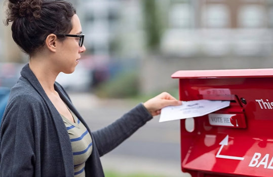 Side profile of a woman placing a mail ballot inside of a ballot drop box.