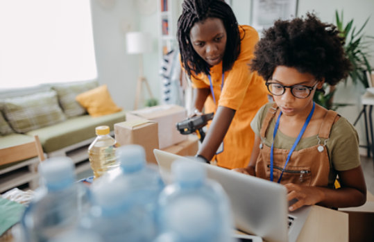 An adult looks over the shoulder of a teenage girl using a laptop in a casual indoor environment.