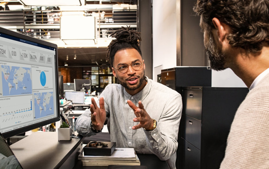 Two colleagues in a modern office discuss analytics displayed on a large monitor with maps, charts, and metrics.