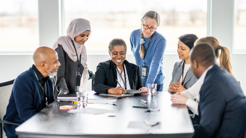 A small team of business professionals huddle around the end of a boardroom table as they look closely at a colleague’s presented ideas.