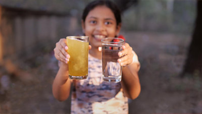 smiling girl holds two glasses of water – one a murky tan and the other crystal clear.