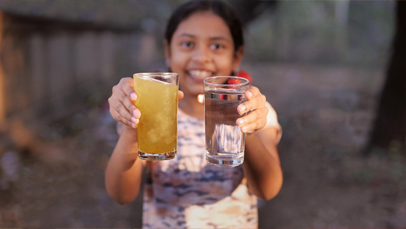 smiling girl holds two glasses of water – one a murky tan and the other crystal clear.