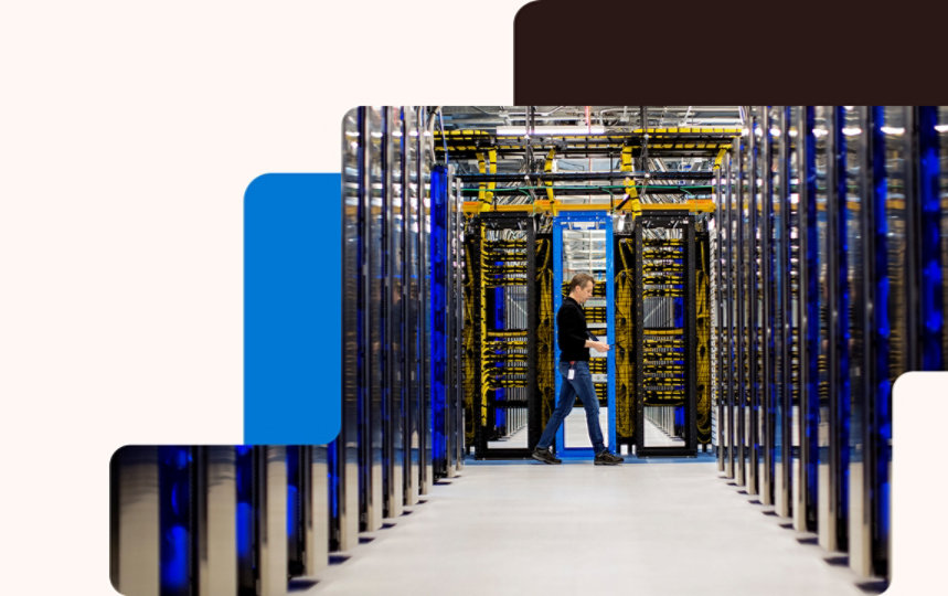A datacenter employee holding a laptop walks along a pathway lined with server stacks.