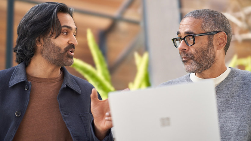 Two men are engaged in a conversation, one holding a laptop.