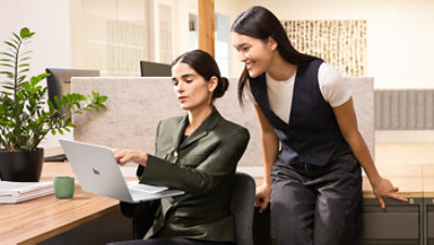 Two female executives in an office. One executive is standing and looking over the shoulder of the second seated executive, holding a Surface Laptop 13.