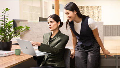Two female executives in an office. One executive is standing and looking over the shoulder of the second seated executive, holding a Surface Laptop 13.