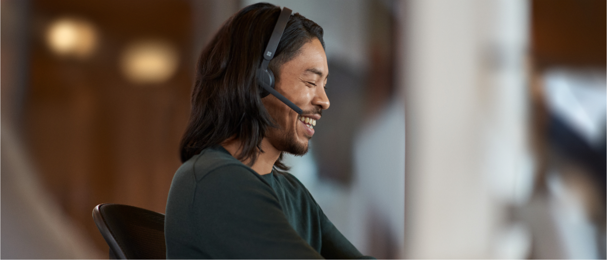 Asian man wearing a headset and smiling while working at a computer in a modern office.