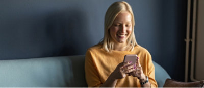 Ženska v rumenem puloverju se smehlja, medtem ko gleda v svoj pametni telefon in sedi na modrem kavču.