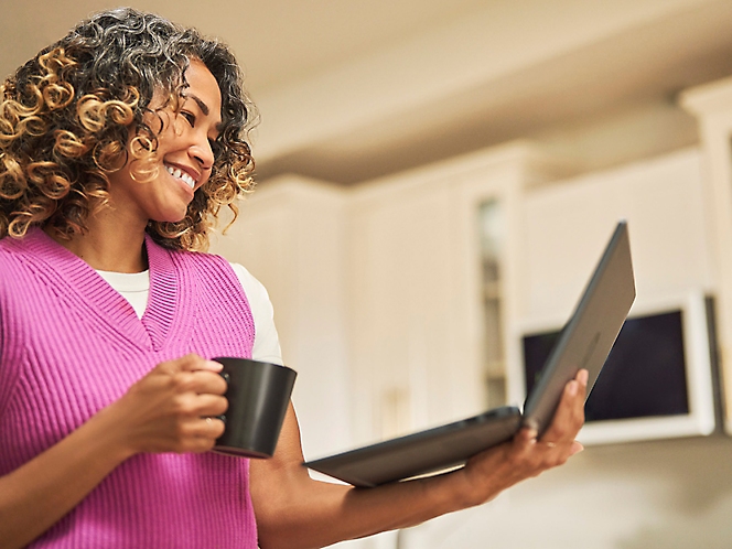 En kvinde smiler og drikker kaffe, mens hun holder en bærbar computer i sin hånd