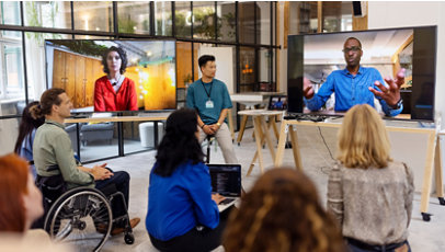 A group of people listen to an on-screen speaker as they face two large displays and a moderator.
