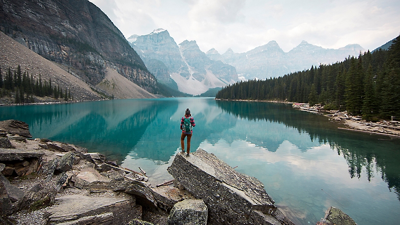 A scenic view of a person standing in front of a landscape of mountains and a crystal blue lake