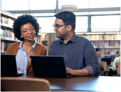 Două persoane stând într-o bibliotecă utilizând laptopuri și purtând o conversație.